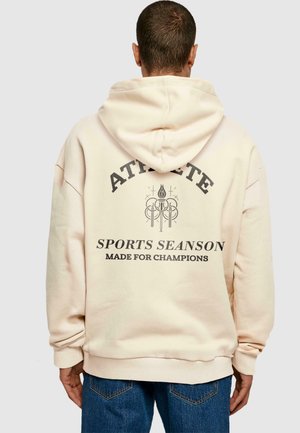 Sudadera crema con un gran gráfico en la parte posterior que lleva los textos "ATLETA", "TEMPORADA DEPORTIVA" y "HECHA PARA CAMPEONES". Textura suave, corte relajado.