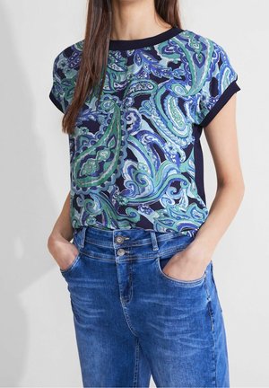 Frau trägt ein kurzärmeliges Top mit blau-grünem Paisley-Muster, in hoch taillierte blaue Jeans gesteckt, mit den Händen in den Taschen.