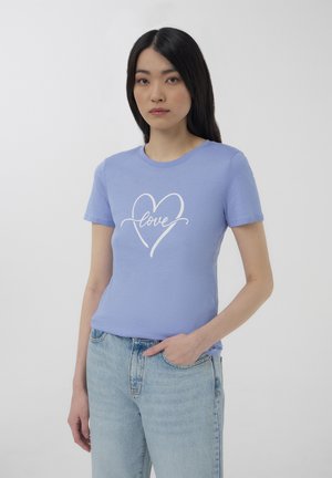 Giovane donna in t-shirt azzurra con scritta "love" e jeans di denim chiaro, in piedi con una mano in tasca su uno sfondo bianco.