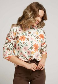 Pull en tricot floral crème, avec des motifs orange, rose et vert. Manches longues avec des poignets côtelés, associé à un pantalon marron.