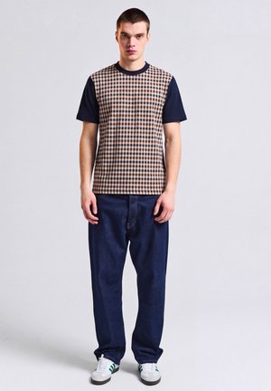 Jonge man draagt een beige en marineblauw geruit overhemd met korte mouwen, donkerblauwe spijkerbroek en witte sneakers, staand tegen een effen lichte achtergrond.