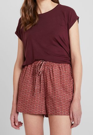 Vrouw draagt een bordeauxrode top met korte mouwen en rode korte broek met patroon en koordje tegen een effen lichte achtergrond.