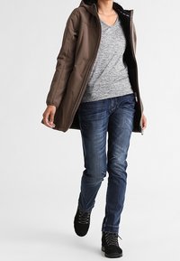 Veste marron à capuche avec une fermeture éclair et une doublure en polaire. Portée par-dessus un t-shirt gris chiné et un jean bleu. Bottes noires à lacets.