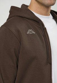 Homme portant un sweat à capuche marron avec un logo blanc représentant deux figures assises sur la poitrine, par-dessus un t-shirt blanc.
