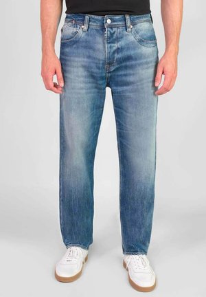 Jean en denim bleu clair à coupe droite, cinq poches et finition délavée. Associés à des baskets blanches avec des semelles en caoutchouc marron.