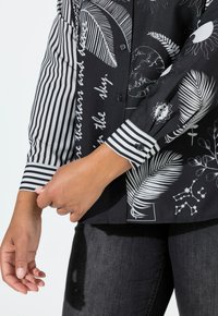 Sort mønstret skjorte med en blanding af hvide blomster- og himmelske motiver på den ene side, kontrasterende med en sort-hvid stribet ærme.