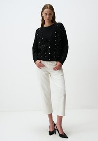 Zwarte kabelgebreide cardigan met parelaccenten, V-halsontwerp en sluiting met vijf knopen, gecombineerd met witte cropped broek en zwarte hakken.