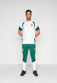 Bielo-zelený športový outfit so zlatými akcentmi a logom. Nohavice sú zelené s bielymi lemy. Oblečené s čiernymi športovými topánkami.