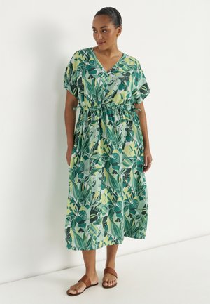 Kobieta stojąca w zielono-żółtej sukience midi w liściasty wzór, z krótkimi rękawami i brązowych płaskich sandałach na białym tle.