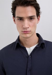 Navyblauw overhemd met knopen en een kraag, met een zachte textuur en een wit ondershirt zichtbaar bij de halslijn. Eenvoudig ontwerp, zonder patronen.