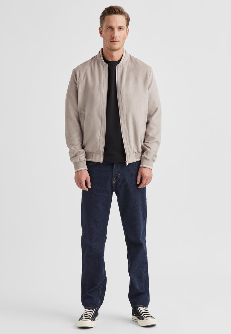 Hellgraue Bomberjacke mit glatter Wildlederstruktur, gerippten Bündchen, Reißverschluss, kombiniert mit dunkelblauen Jeans und schwarzen Turnschuhen.