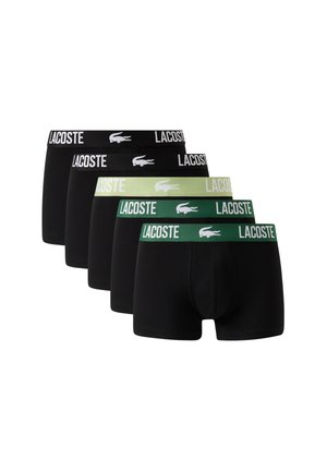 Мъж, облечен с черни боксерки Lacoste с зелен колан, стоящ бос на бял фон.