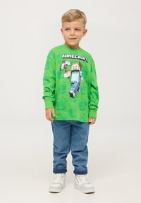 Maglietta verde a maniche lunghe con grafica di un personaggio di Minecraft sul davanti, abbinata a jeans in denim blu e sneakers bianche.