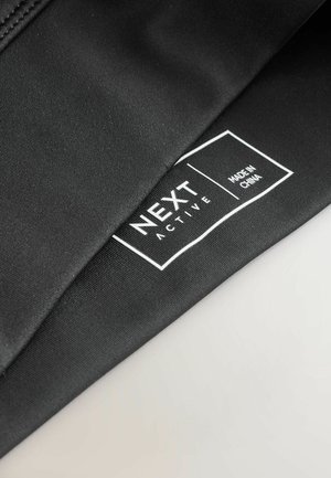 Juodos spalvos sportinis audinys su lygiu paviršiumi turi baltą etiketę, kurioje parašyta „NEXT ACTIVE“ ir „MADE IN CHINA“ stačiakampio formos rėmo viduje.