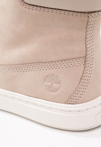 Beige suede ankle boot featuring stitched detailing, a white rubber sole, and an embossed tree logo on the outer side.