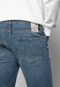 Blå denim jeans med hög midja, med två bakfickor och ett skötselband. Sömmarna är i en kontrasterande guldnyans.