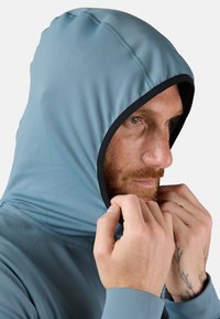 Lichtblauwe capuchontrui met een zwarte binnenvoering; heeft een gladde textuur en een aansluitend design, met handen die de capuchon vastgrijpen.