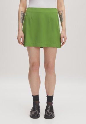 Groene, knielange rok van gladde stof met een lichte glans. Bevat twee voorplooien en een nette zoom. Draagt bij zwarte schoenen en sokken.