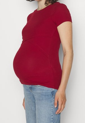 Chemise de maternité rouge à la coupe ajustée, manches courtes, tissu texturé, associée à un jean bleu clair. Ventre de grossesse visible.