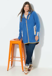 Blaue Fleecejacke mit einem orangefarbenen Reißverschluss und Saum, mit Seitentaschen. Kombiniert mit dunklen Jeans und mehrfarbigen Sneakern. Orangenes Hocker im Hintergrund.
