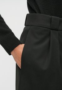 Μαύρο ταγέρ παντελόνι με φαρδύ λάστιχο στη μέση, που διαθέτει πιέτες και πλαϊνές τσέπες, συνδυασμένο με πλεκτό μαύρο μανίκι.