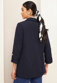 Blazer sartorial navy con maniche arrotolate e dettagli in oro, abbinato a jeans. Presenta una texture liscia e un design strutturato.