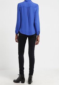 Blå långärmad blus med en ryggplatta och veckdetaljer, kombinerad med figurnära svarta jeans och stövlar med ankeldesign.