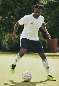Camisa blanca de manga corta con el logo negro de Adidas, pantalones cortos negros y calcetines blancos con rayas negras. Botas de fútbol verdes en acción cerca de un balón de fútbol colorido.