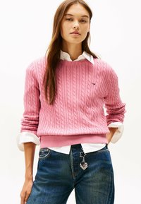 Pullover in maglia rosa con orlo a coste, indossato sopra una camicia bianca con colletto. Jeans in denim con charm decorativi attaccati alla tasca.