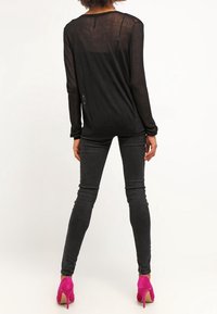 Svart genomskinlig långärmad topp bärs med mörka slim-fit jeans och klarröda högklackade skor, vilket betonar form och materialkontrast.