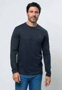 Homme aux cheveux foncés et barbe, portant une chemise à manches longues bleu marine et un jean bleu, debout devant un fond clair uni.