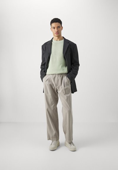 Svart blazer over en lys grønn ribbestrikket genser, kombinert med beige plissert bukser og hvite sneakers mot en nøytral bakgrunn.