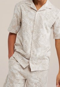 Svetle béžová košile s krátkým rukávem a odpovídajícími šortkami s bílými květinovými vzory, s klopovým límcem a zapínáním na knoflíky.
