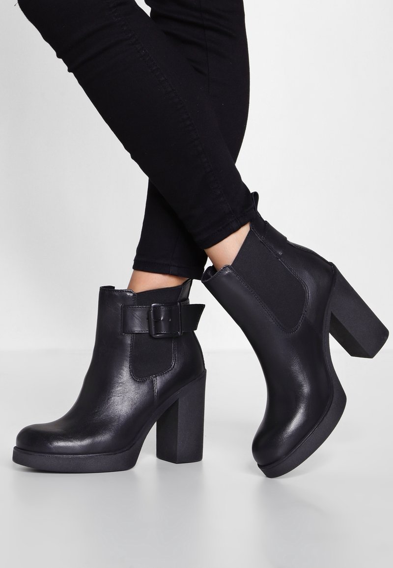 Bottes courtes en cuir noir avec talons épais et panneaux latéraux élastiques, portées avec un pantalon noir ajusté sur un fond uni.