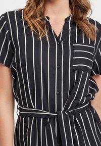 Vestido negro con rayas verticales blancas, con botones en la parte delantera, mangas cortas, cuello mao y cintura ajustada con un lazo. Presenta un bolsillo en el pecho.