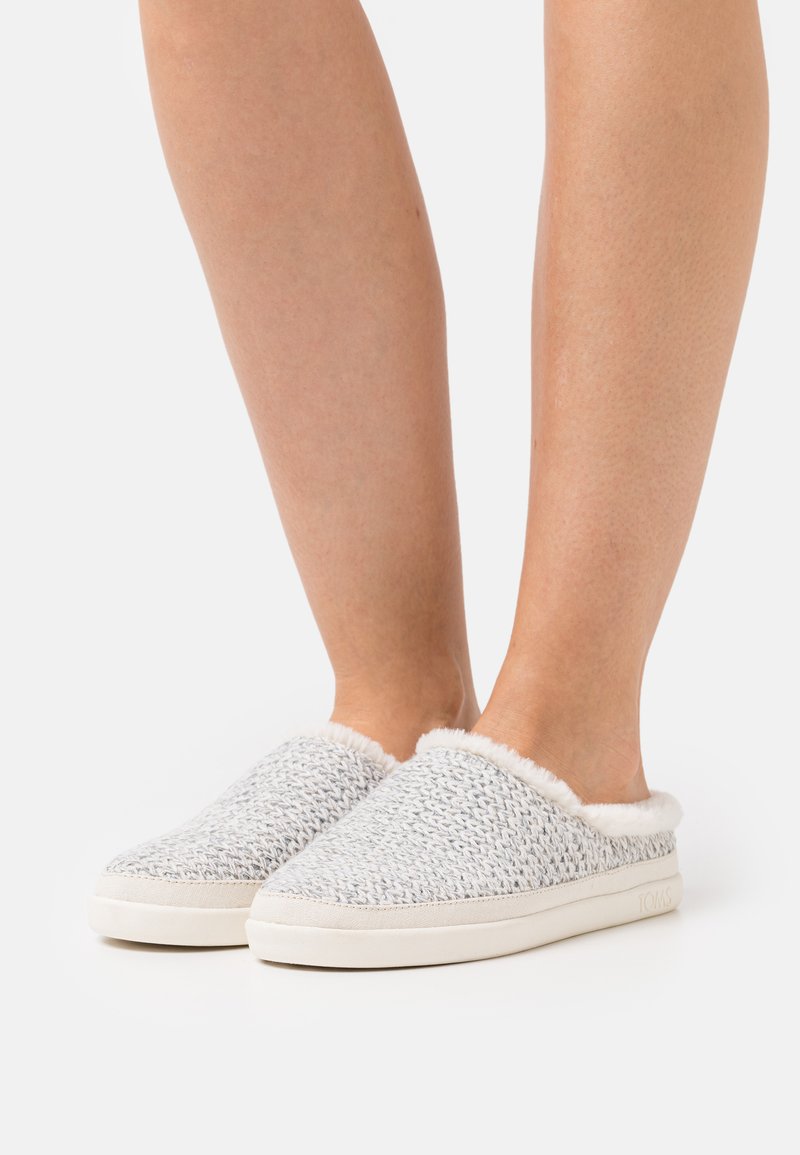 Piedi che indossano pantofole slip-on grigio chiaro in maglia con fodera in pelliccia bianca e suole in gomma bianca su uno sfondo neutro.