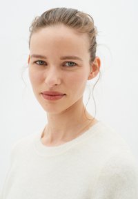 Pulover bej deschis cu o textură moale și guler rotund. Modelul are părul drept, ochi albastru-verzui și o expresie neutră pe un fundal alb.