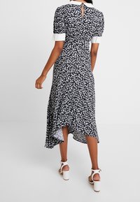 Robe marine avec imprimé papillon, manches courtes bouffantes et dos avec ouverture en forme de goutte. Ourlet asymétrique et tissu léger. Portée avec des talons blancs.