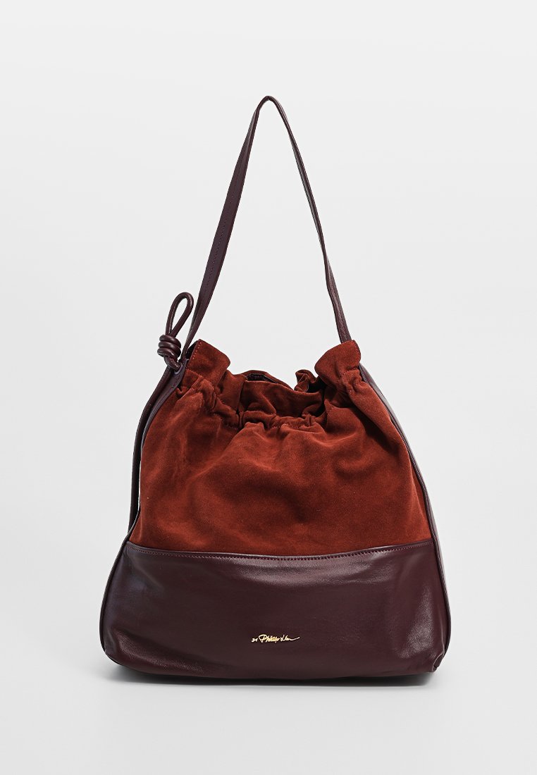 Brązowa skórzana torba typu bucket z zamszowymi detalami, ze ściąganym wierzchem, pojedynczym paskiem na ramię oraz eleganckim złotym akcentem logo na dole.