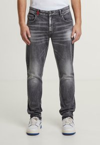 Jeans de mezclilla negros con un lavado desgastado, diseño de cinco bolsillos y acento rojo en la cintura. Combinados con zapatillas blancas con detalles en azul.