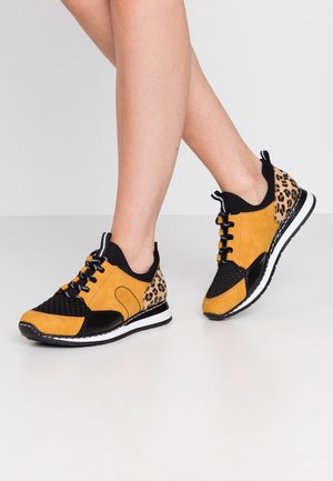 Osoba nosząca musztardowo-żółte, czarne i w panterkę sneakersy, stojąca ze skrzyżowanymi nogami na jasnoszarej podłodze i tle.
