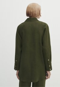 Giacca over-size in lino verde scuro con colletto a punta, maniche lunghe e polsini con bottoni, caratterizzata da un design con schiena dritta.