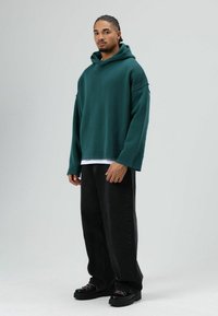 Grüner übergroßer Hoodie mit Kapuze, gerippten Bündchen und rohem Saum, kombiniert mit lockeren schwarzen Jeans und klobigen schwarzen Schnürschuhen.