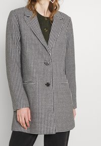 Manteau à motif pied-de-poule noir et blanc avec une coupe structurée, deux boutons, revers crantés et poches latérales, porté sur un haut vert.