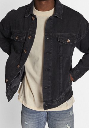 Mężczyzna w czarnej, rozpiętej kurtce jeansowej na beżowej koszulce oraz podniszczonych niebieskich dżinsach, z rękami w kieszeniach kurtki.