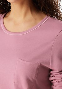Camicia a maniche lunghe rosa con scollatura a girocollo, dotata di taschino sul lato sinistro e dettagli arricciati sulle maniche. Tessuto morbido.