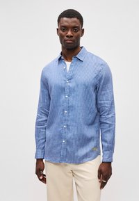 Μπλε ριγέ πουκάμισο με μακριά μανίκι α και γιακά με κουμπί, φτιαγμένο από ελαφρύ υλικό, με μια διακριτική λεπτομέρεια λογότυπου στο στρίφωμα.