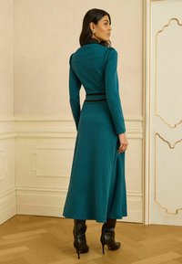 Rochie tricotată verde albastruie cu corset fixat, accente negre la talie și mâneci lungi, asortată cu cizme înalte negre cu toc. Podea texturată.