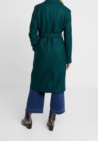 Manteau en laine teal avec une taille ceinturée, des manches longues et un col. Associé à un pantalon en denim à jambes larges et des bottines à imprimé serpent.