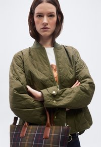 Γυναίκα με κοντά καστανά μαλλιά, που φοράει αντιολισθητικό μπουφάν ελιάς πάνω από ένα λευκό πουκάμισο με τύπωμα σκύλου, κρατώντας μια καρό τσάντα με καφέ δερμάτινα χερούλια.